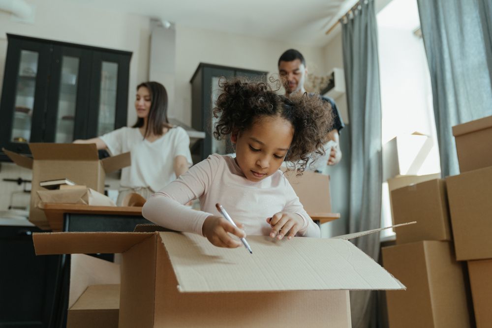 A girl in white long sleeve shirt writing on a cardboard box during a family move.